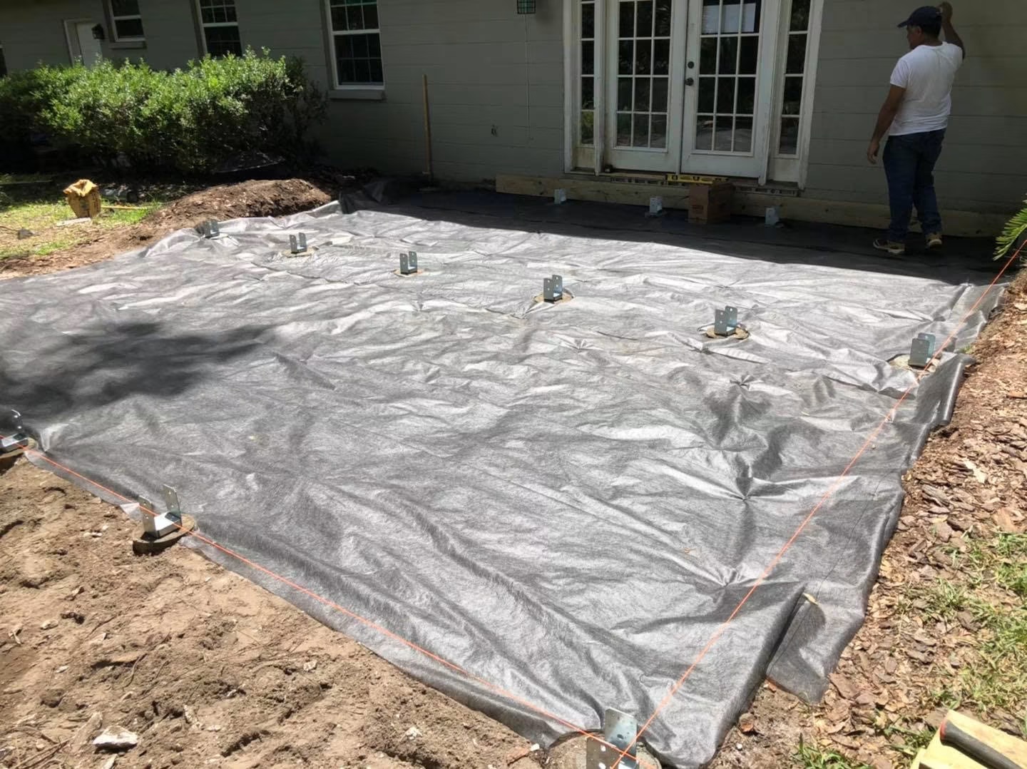 Deck build step 1 of 4: the leveled dirt footprint covered in black weed-barrier fabric with galvanized metal post bases set into concrete footings